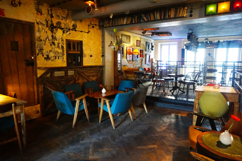 Jozef K cafe in Gdansk, Poland. Several mismatched chairs and tables are spread out with railings and vintage furniture and decor at the back.