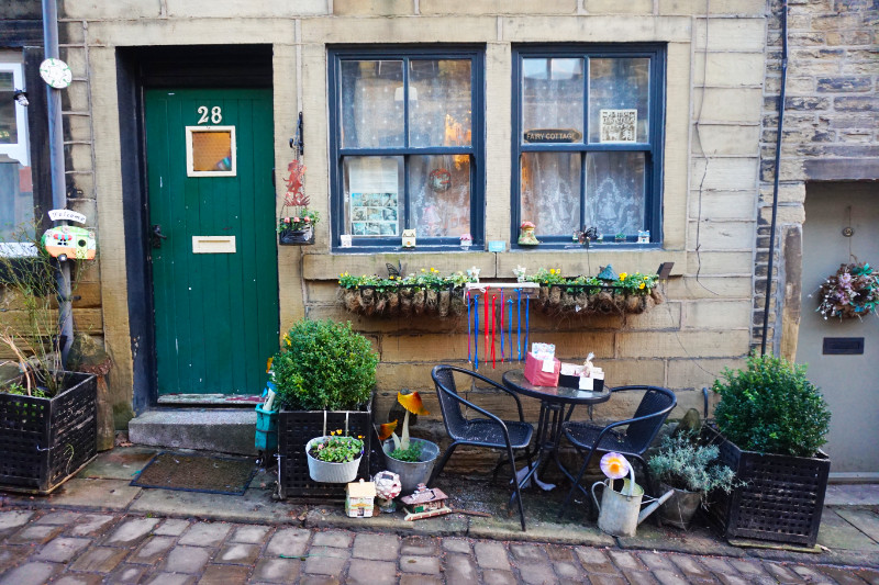 House in Haworth, Yorkshire. On the left is a green door, with two windows to the right with decorations and signs. A table and chairs are outside with plant pots and watering cans.