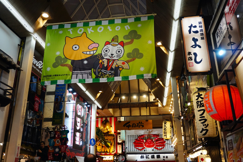 Dotonbori, Osaka, Japan. It's a covered walkway with a mascot advert for Tokoton, restaurant signs, and a big crab at the end.
