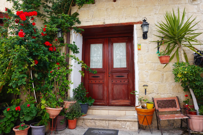 Pretty house in Omodos, Cyprus. A brown double door is surrounded by plans, with pots and bushes on one side and hanging baskets and tropical plants on the right.