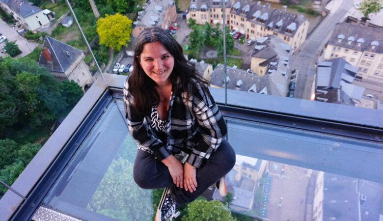The viewpoint at the Pfaffenthall elevator, Luxembourg. I am sitting on a glass floor wearing black jeans and a black and white checked shirt, looking up at the camera.