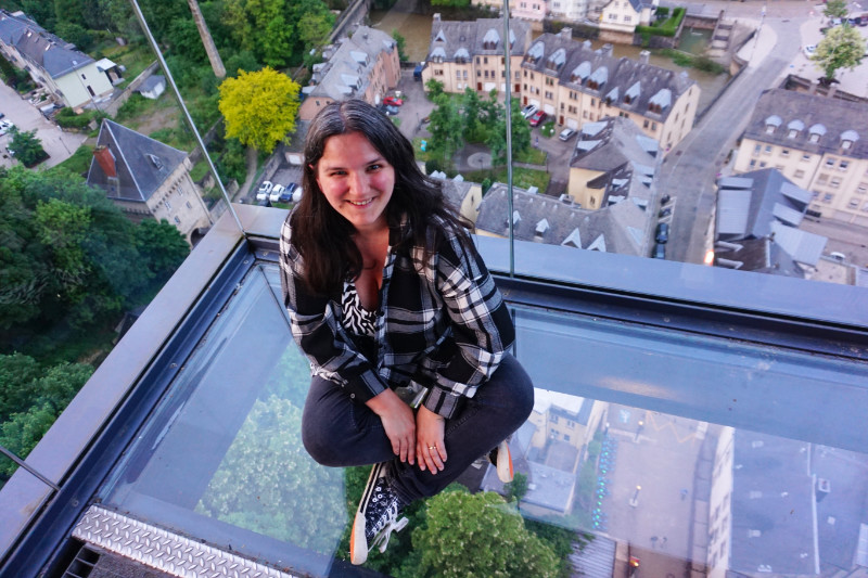 The viewpoint at the Pfaffenthall elevator, Luxembourg. I am sitting on a glass floor wearing black jeans and a black and white checked shirt, looking up at the camera.