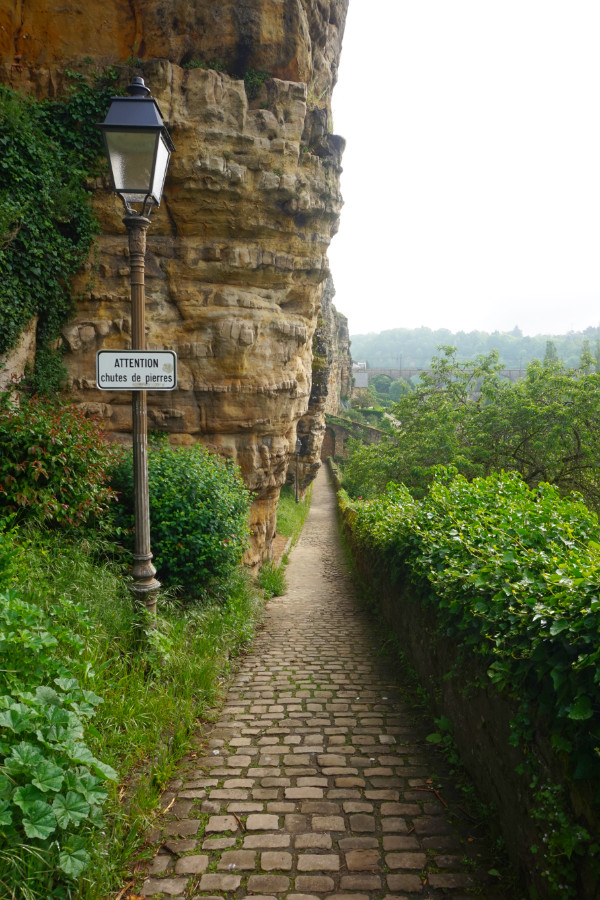 Wenzel Circular Walk, Luxembourg. A very narrow path leads alongside the bottom of the cliff on one side, with bushes on the other. In the foreground on the left is an old street lamp.