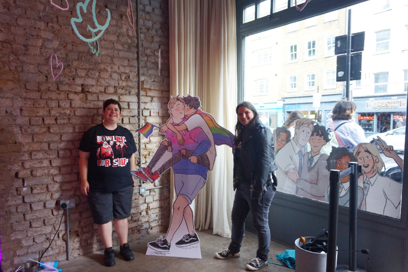 Heartstopper pop up in Shoreditch, London. There is a cardboard cut-out of Nick giving Charlie a piggy back. Charlie is wearing a rainbow cape and holding a rainbow flag. My friend and I are standing either side. In the window to the right of me is a decal of all the main characters from the show. Above are coloured leaves hanging from the ceiling.
