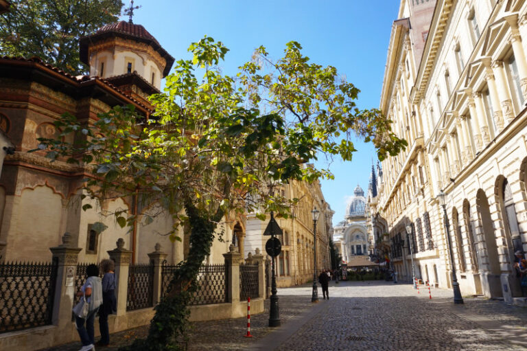 Strada Stavropoleos, Bucharest, Romania. A monastery is on the left, and a tree leaning into the street from next to the monastery. At the end is a huge, beautiful building with a glass dome roof.
