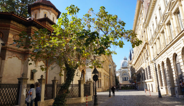 Strada Stavropoleos, Bucharest, Romania. A monastery is on the left, and a tree leaning into the street from next to the monastery. At the end is a huge, beautiful building with a glass dome roof.
