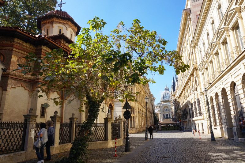 Strada Stavropoleos, Bucharest, Romania. A monastery is on the left, and a tree leaning into the street from next to the monastery. At the end is a huge, beautiful building with a glass dome roof.