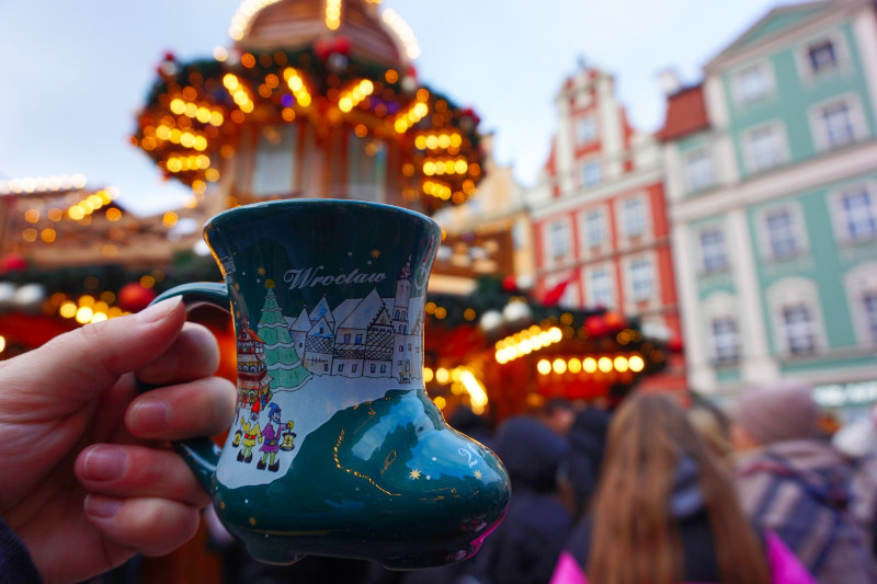 Holding a cup at the Christmas markets, Wroclaw, Poland. It's a green cup shaped like a boot and has a snowy scene on it saying "Wroclaw". Behind it is a lit-up round stall and a couple of pretty, colourful buildings, out of focus.