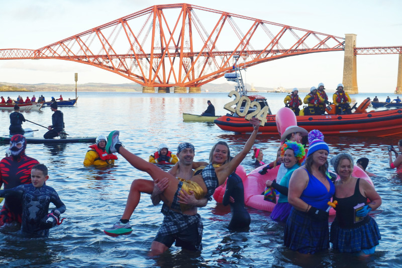 Loony Dook, South Queensferry, Scotland. Several people are in the water in front of the Forth Bridge. A man is holding a woman, and she's holding a sign saying 2024. Some ladies next to them are posing for a photo. There's an inflatable pink flamingo and people dressed as superheroes, and behind them is a lifeboat with real superheroes, and some other boats.